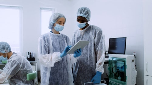两名身穿防护装备的医务人员正看着平板电脑，确保质量管理。